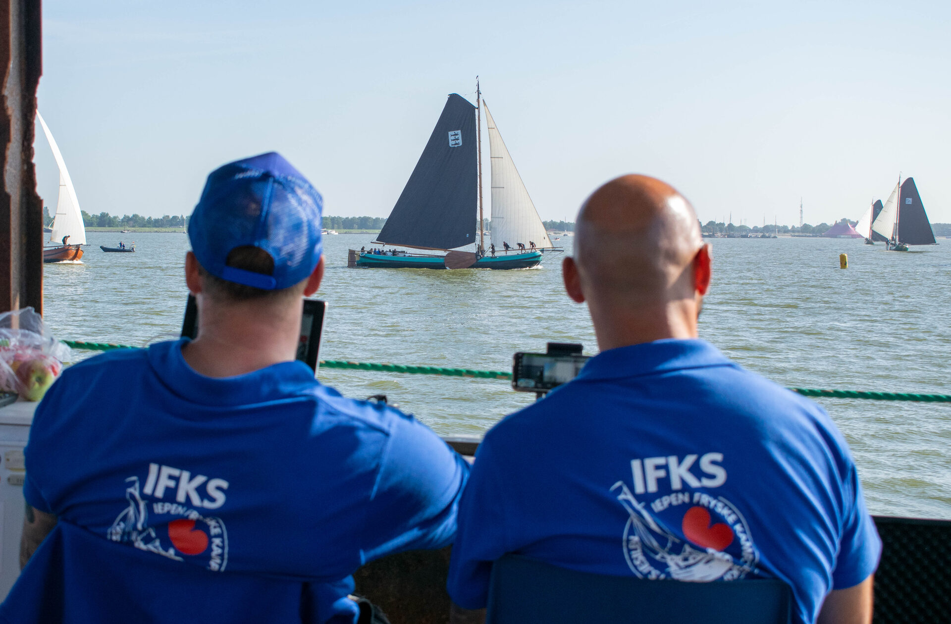 Twee crewleden van IFKS TV kijken vanaf het dek naar een passerend skûtsje